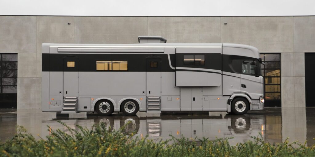 Extended luxury Scania motorhome with multiple axles and side living module, shown in profile outside a contemporary building