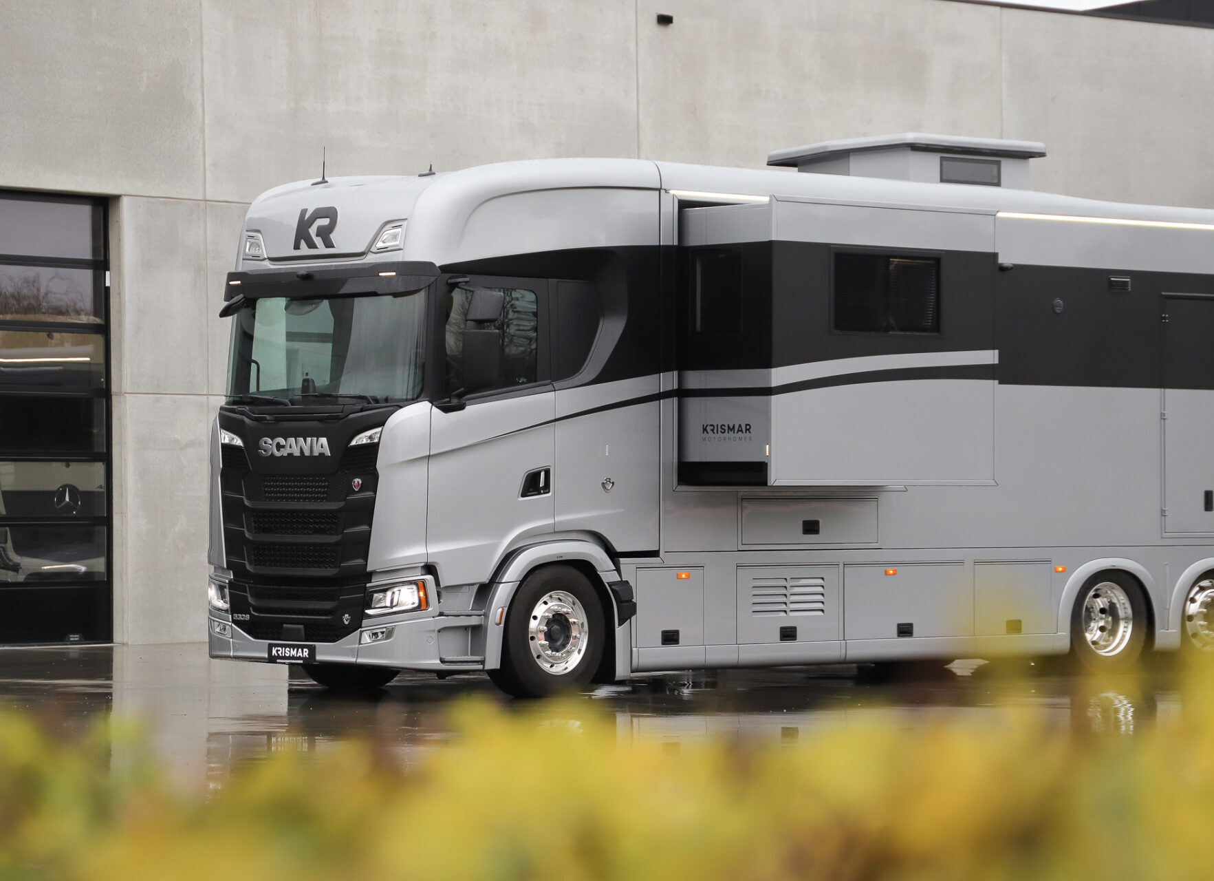 Large luxury motorhome built on a Scania truck chassis, finished in silver and black, parked in front of a modern industrial building