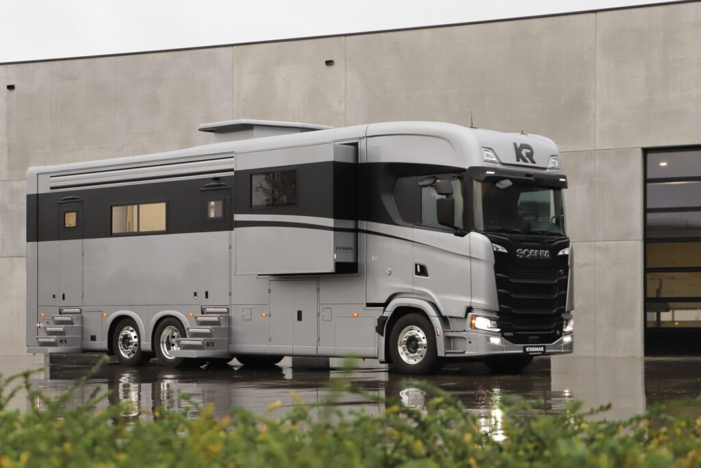 High-end Scania-based motorhome with extended side module and sleek grey exterior, parked in front of a contemporary industrial building